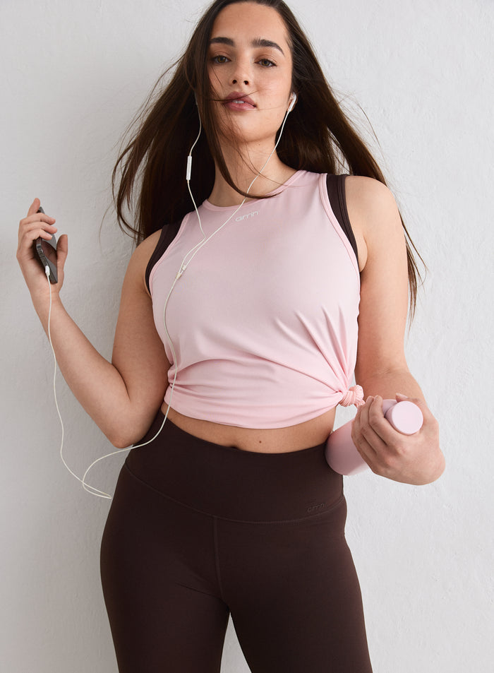 A woman wearing a pink aim'n Soft Basic Singlet tied at the waist, paired with brown leggings and holding a phone and water bottle [L-174cm]