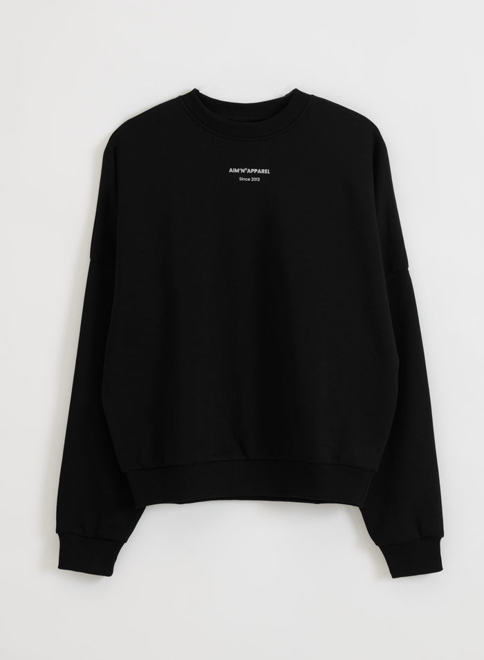 Relaxed black terry sweatshirt with a silver aim'n logo, ribbed hem, and cuffs, shown in a flatlay.