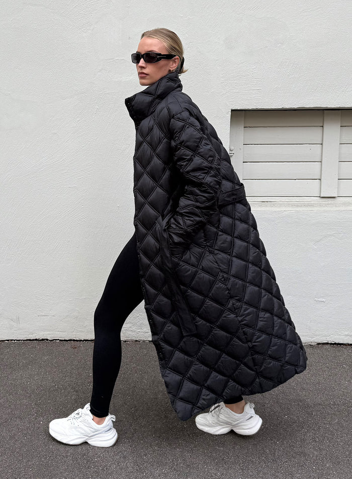 Long black diamond-quilted coat with a high collar worn by a woman walking outdoors. [M-175cm]