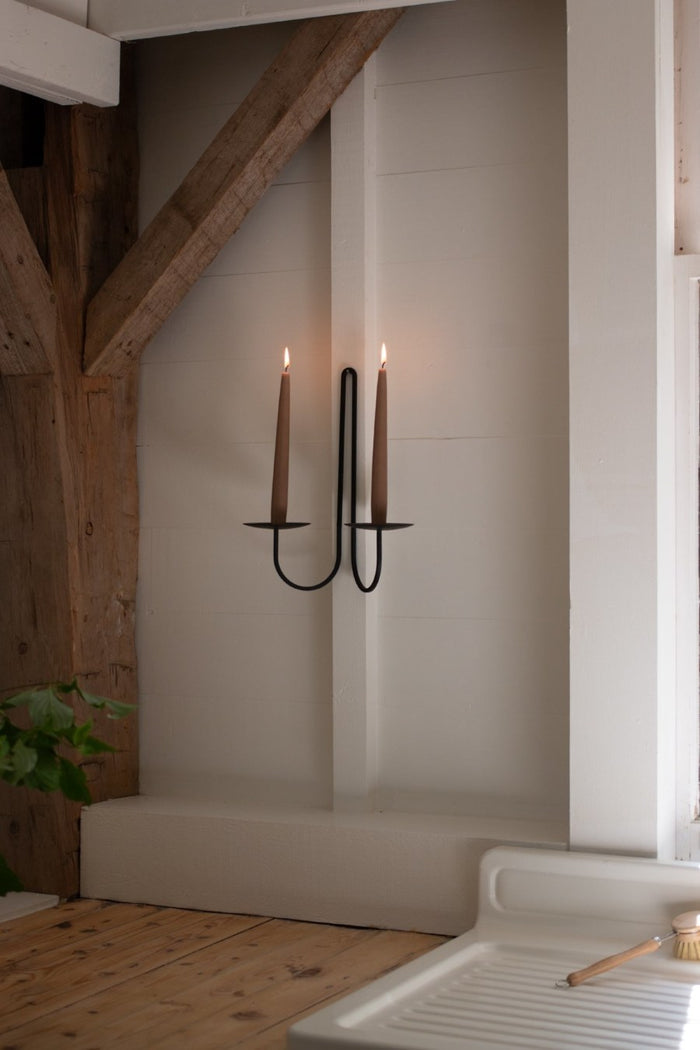 black iron sconce with two candles on a white wall in a room with wooden beams and a plant.