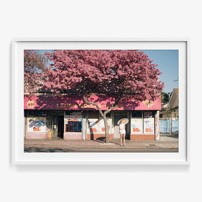 Photograph in a thin white frame showing a woman with a parasol standing by a pink building under a flowering tree