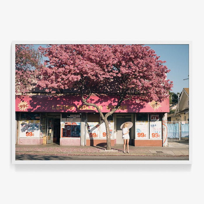 Photograph in a thick white frame showing a woman with a parasol standing by a pink building under a flowering tree