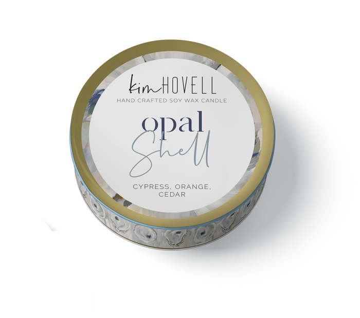A travel tin candle with a label reading 'Kim Howell Hand Crafted Soy Wax Candle Opal Shell, scents of Cypress, Orange, Cedar', with a background pattern of shells.