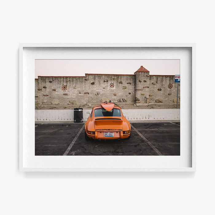 Rear view of an orange Porsche with a surfboard on its roof parked in front of a stone wall framed in white