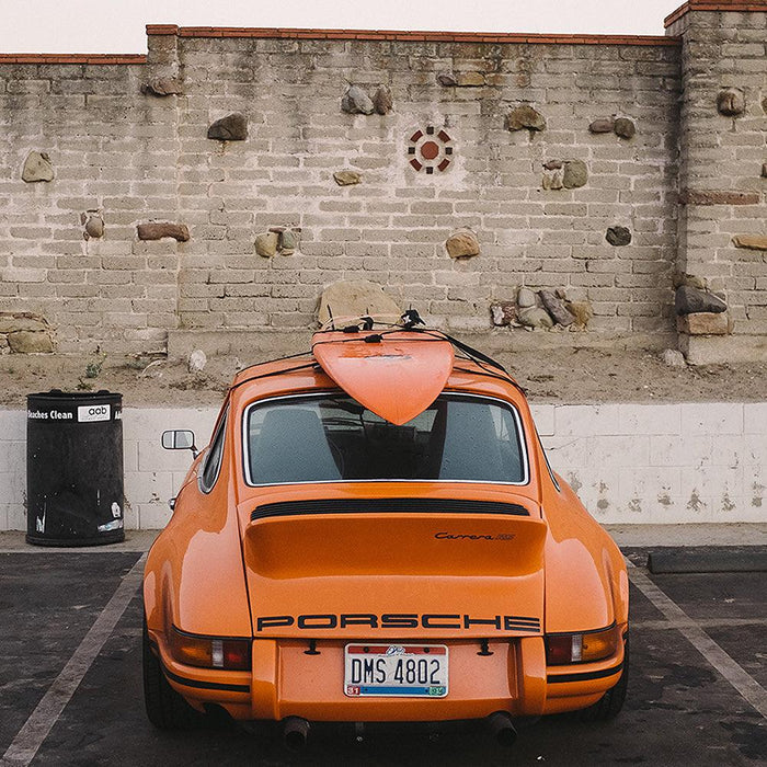 Orange Porsche 911 Carrera 2 7 RS with a surfboard on top parked in front of a stone wall