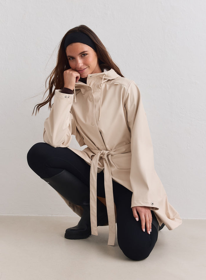 Sand-colored aim'n Rain Jacket with a belted waist and adjustable hood, worn with black leggings. [M-178cm]