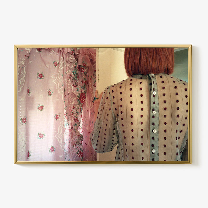 A woman with red hair wearing a sheer polka-dot blouse with button-up back facing pink floral curtains